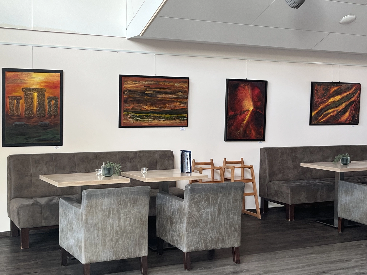 Hannelore Drees-Kiefhaber - Acrylmalerei von der Nordseeküste - Eine moderne Kunstgalerie oder ein Loungebereich mit grau gepolsterten Stühlen, Sitznischen, Holzfußboden, abstrakten Gemälden an der Wand und einzigartigen Deckenleuchten.