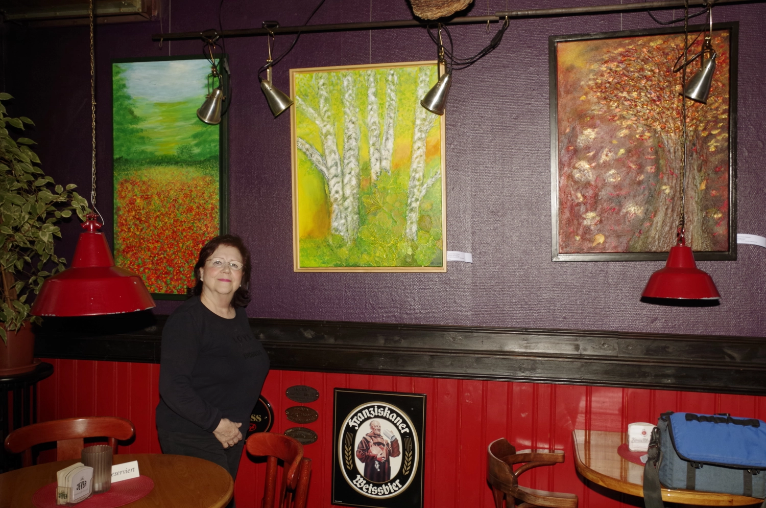 Hannelore Drees-Kiefhaber - Acrylmalerei von der Nordseeküste - Eine Frau in einem schwarzen Hemd steht in einer Kneipe mit lila und roten Wänden, neben drei Gemälden, die Naturlandschaften darstellen, und einem Franziskaner-Bierschild.