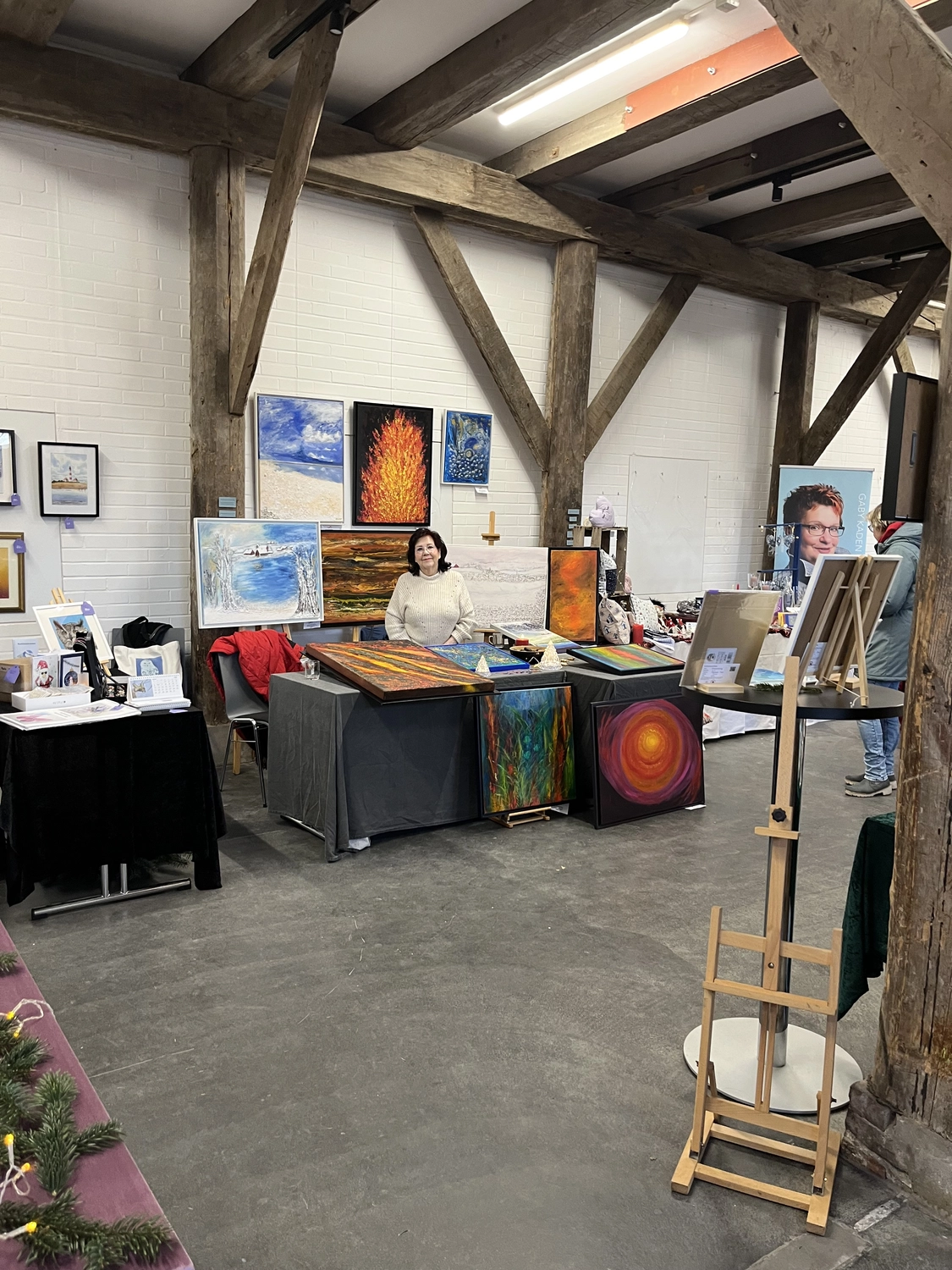 Hannelore Drees-Kiefhaber - Acrylmalerei von der Nordseeküste - Eine Frau sitzt an einem Ausstellungstisch in einer rustikalen Kunstgalerie oder einem Atelier, umgeben von verschiedenen Gemälden und Künstlerbedarf.