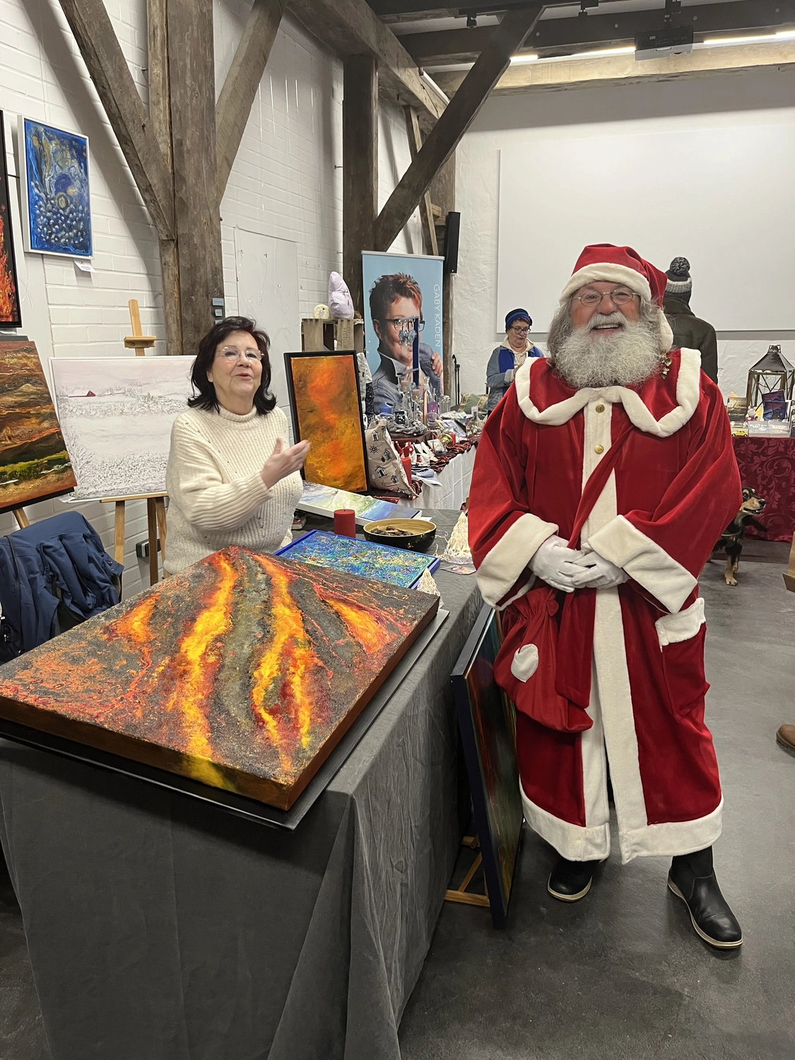 Hannelore Drees-Kiefhaber - Acrylmalerei von der Nordseeküste - Hinter einem Kunsttisch, auf dem ein farbenfrohes, abstraktes Gemälde ausgestellt ist, steht eine Frau neben einem Mann, der als Weihnachtsmann verkleidet ist.