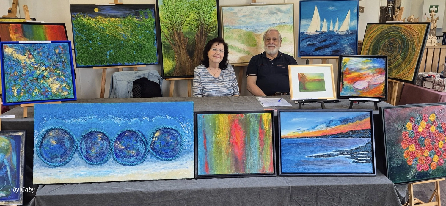 Hannelore Drees-Kiefhaber - Acrylmalerei von der Nordseeküste - Ein Mann und eine Frau sitzen hinter einem Tisch, auf dem viele farbenfrohe Gemälde auf einer Kunstmesse ausgestellt sind.