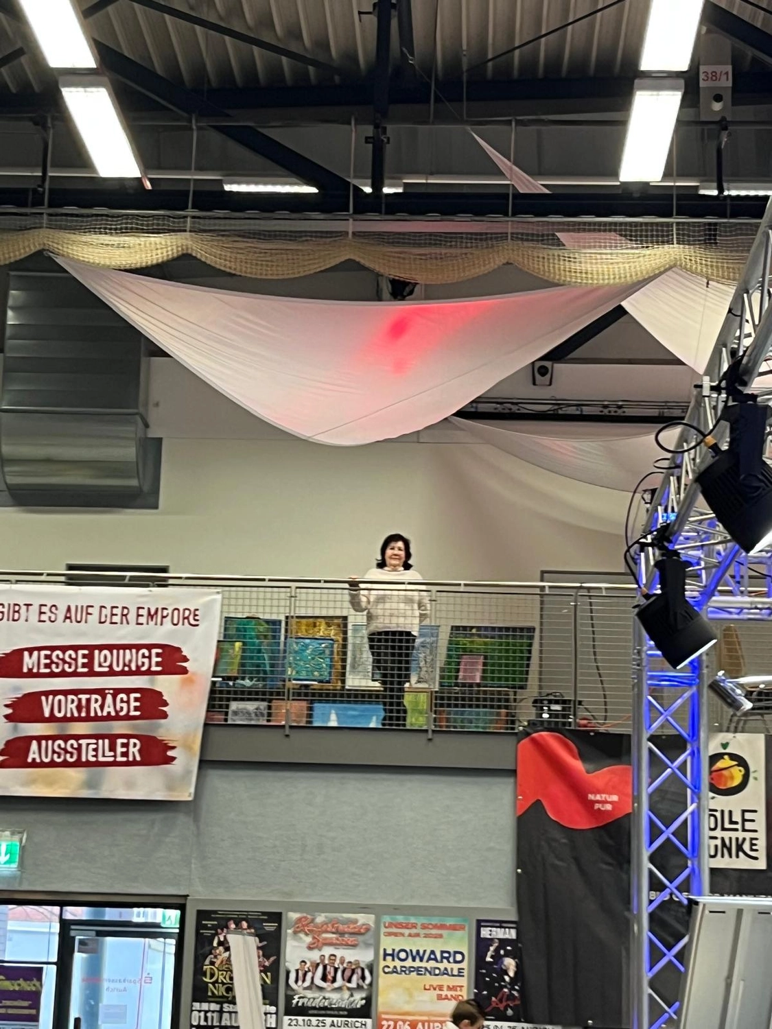 Hannelore Drees-Kiefhaber - Acrylmalerei von der Nordseeküste - Ein Veranstaltungsraum mit Balkon, auf dem eine Person steht. Banner mit deutscher Aufschrift, darunter „MESSE LOUNGE“, „VORTRÄGE“ und „AUSSTELLER“, sind zu sehen. Bühnenbeleuchtung und Plakate sind ebenfalls vorhanden.