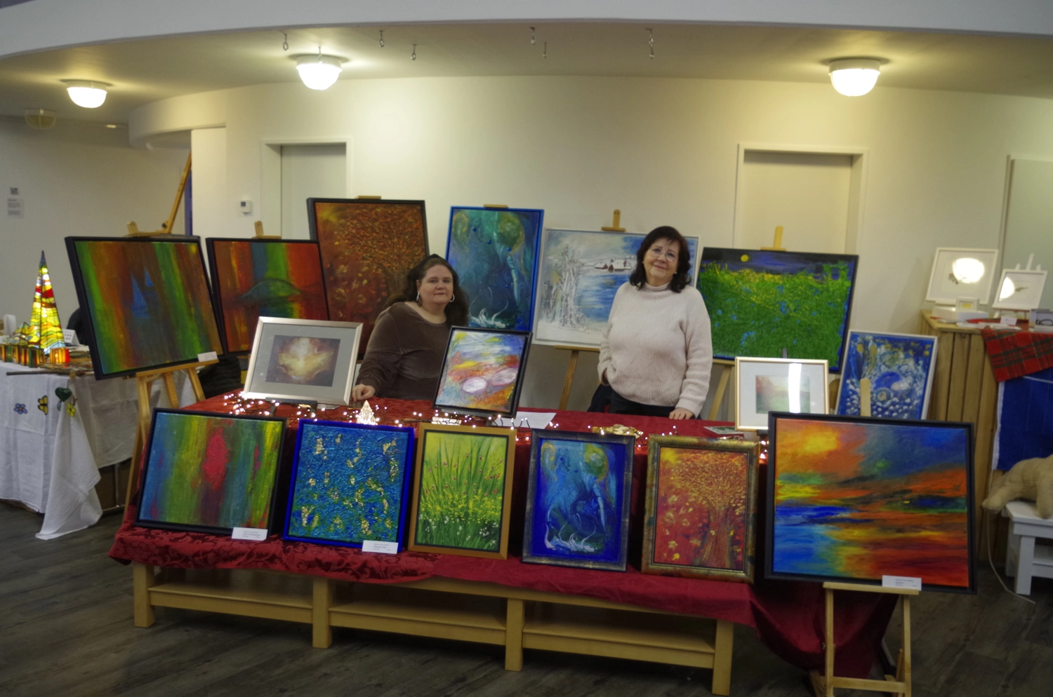 Hannelore Drees-Kiefhaber - Acrylmalerei von der Nordseeküste - Zwei Frauen stehen hinter einem Tisch, auf dem verschiedene farbenfrohe Gemälde bei einer Kunstausstellung ausgestellt sind.