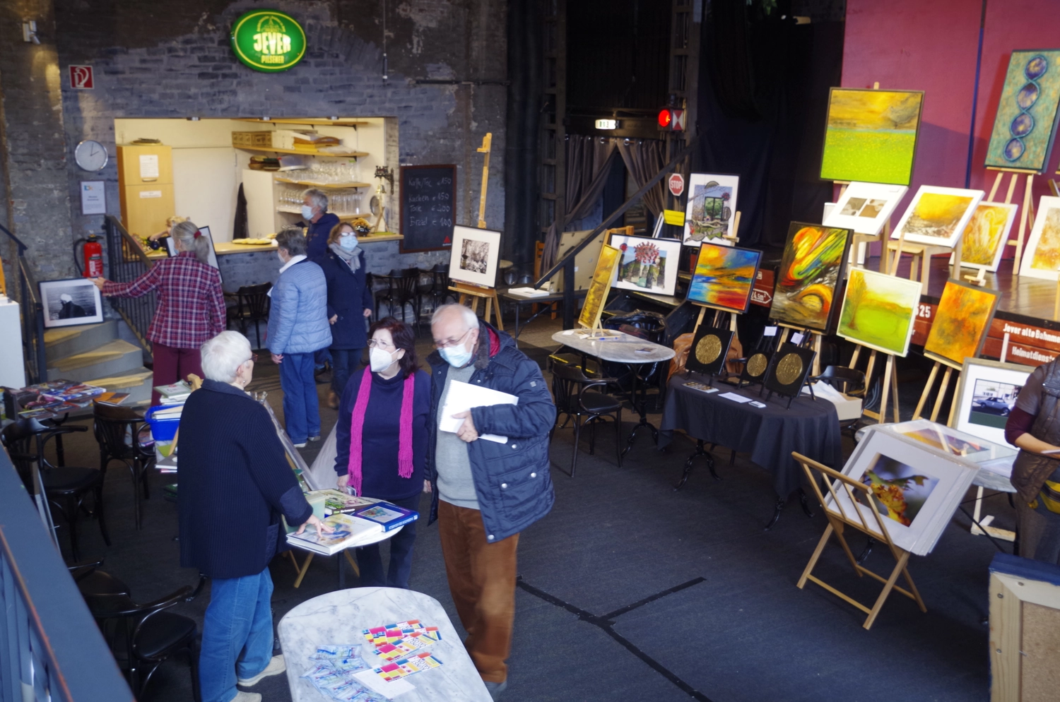 Hannelore Drees-Kiefhaber - Acrylmalerei von der Nordseeküste - Eine Innenaufnahme einer Kunstausstellung oder eines Kunstmarktes, auf der mehrere maskierte Personen verschiedene Gemälde und Kunstwerke betrachten, die auf Staffeleien und Tischen ausgestellt sind. Im Hintergrund ist ein kleines Café zu sehen.