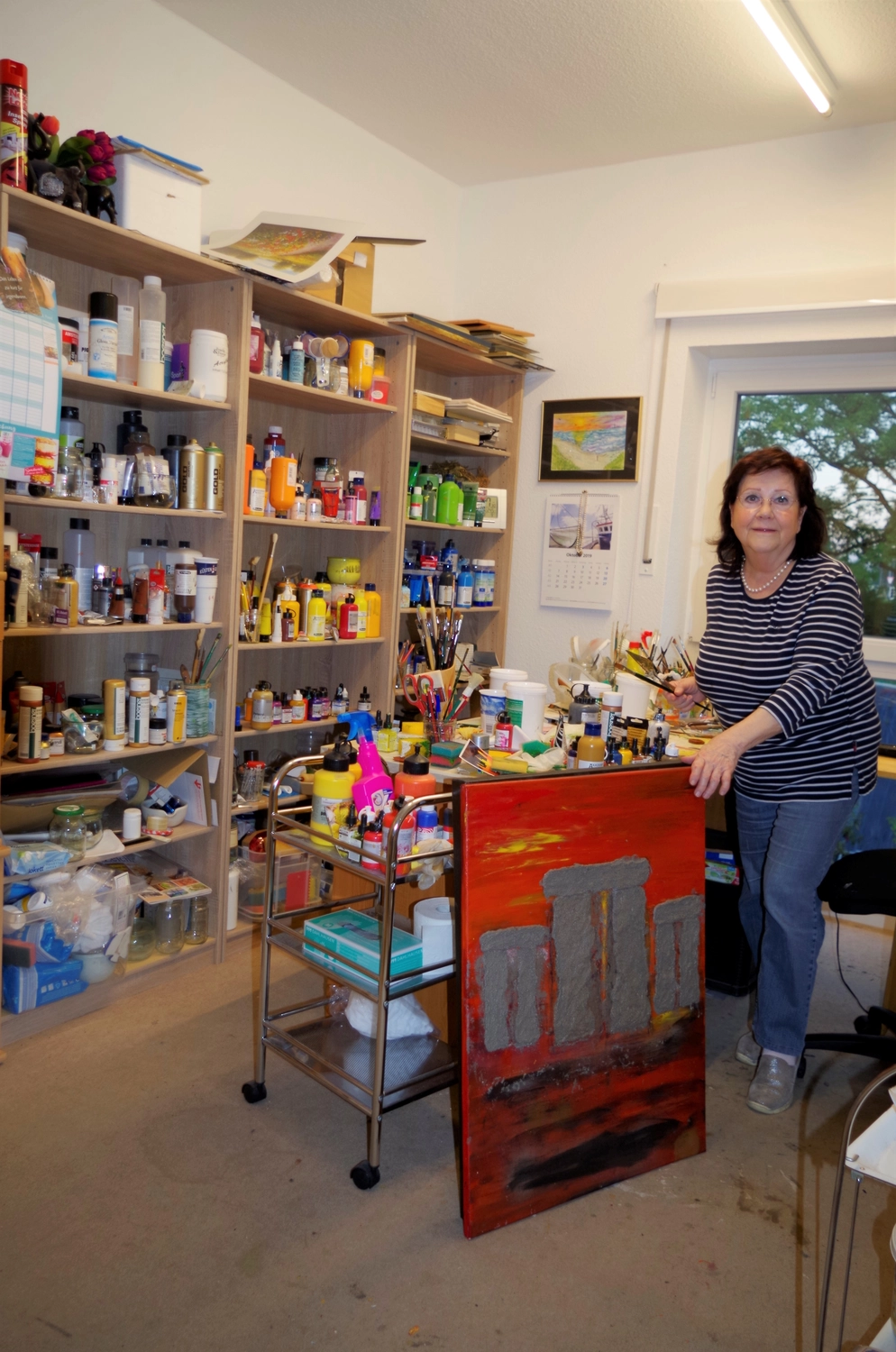 Hannelore Drees-Kiefhaber - Acrylmalerei von der Nordseeküste - Stephanie Drees steht in einem gestreiften Hemd in ihrem Atelier, umgeben von Regalen voller Künstlerbedarf und einem unfertigen Gemälde auf einer Staffelei.